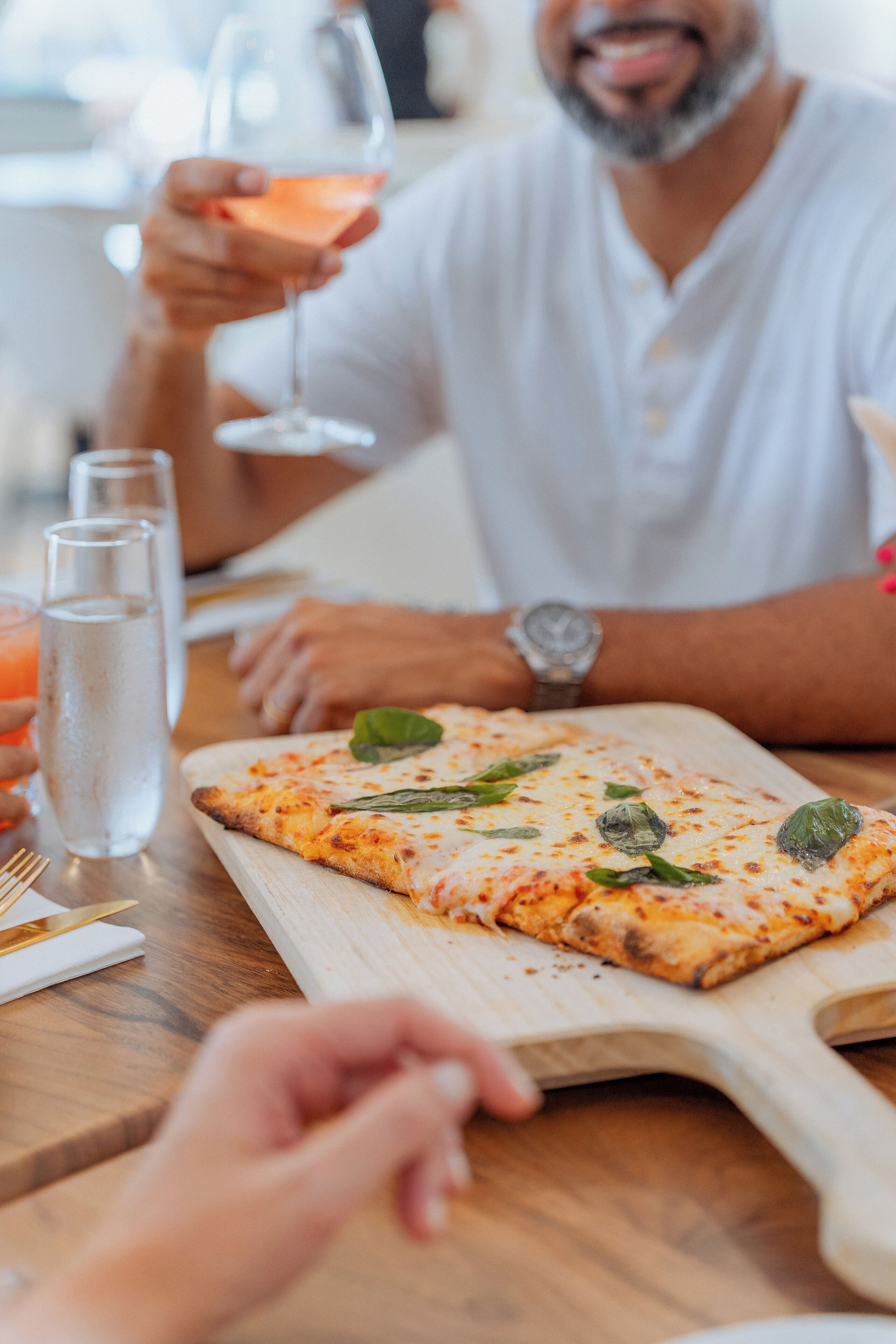 A dining table with a pizza on a wooden board, a glass of water, and a person holding a glass.