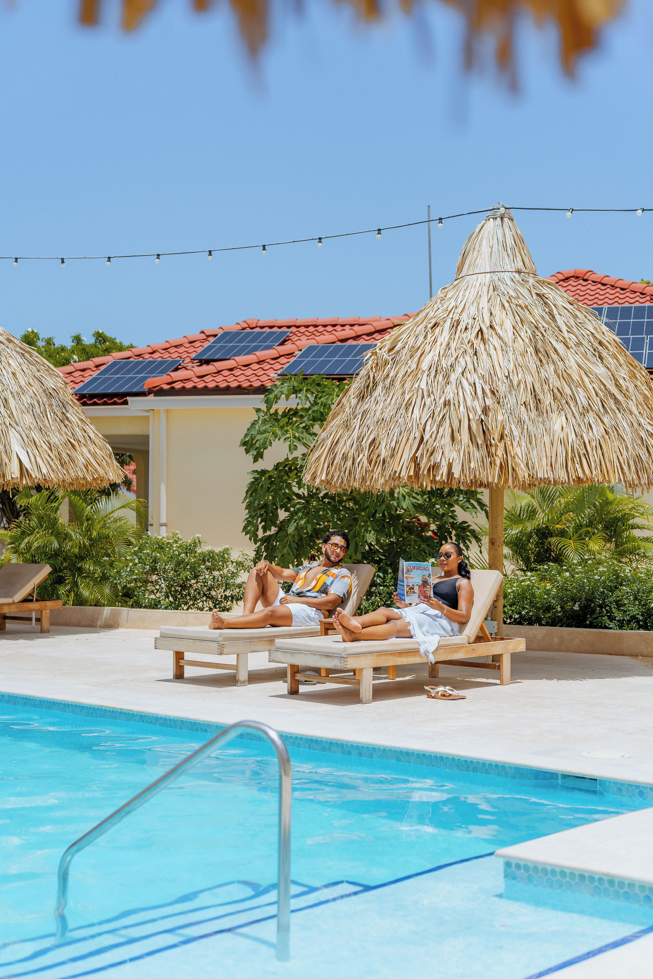 Een zwembadgedeelte met ligbedden en rieten parasols, een heldere lucht en een gebouw met een rode dakbedekking.