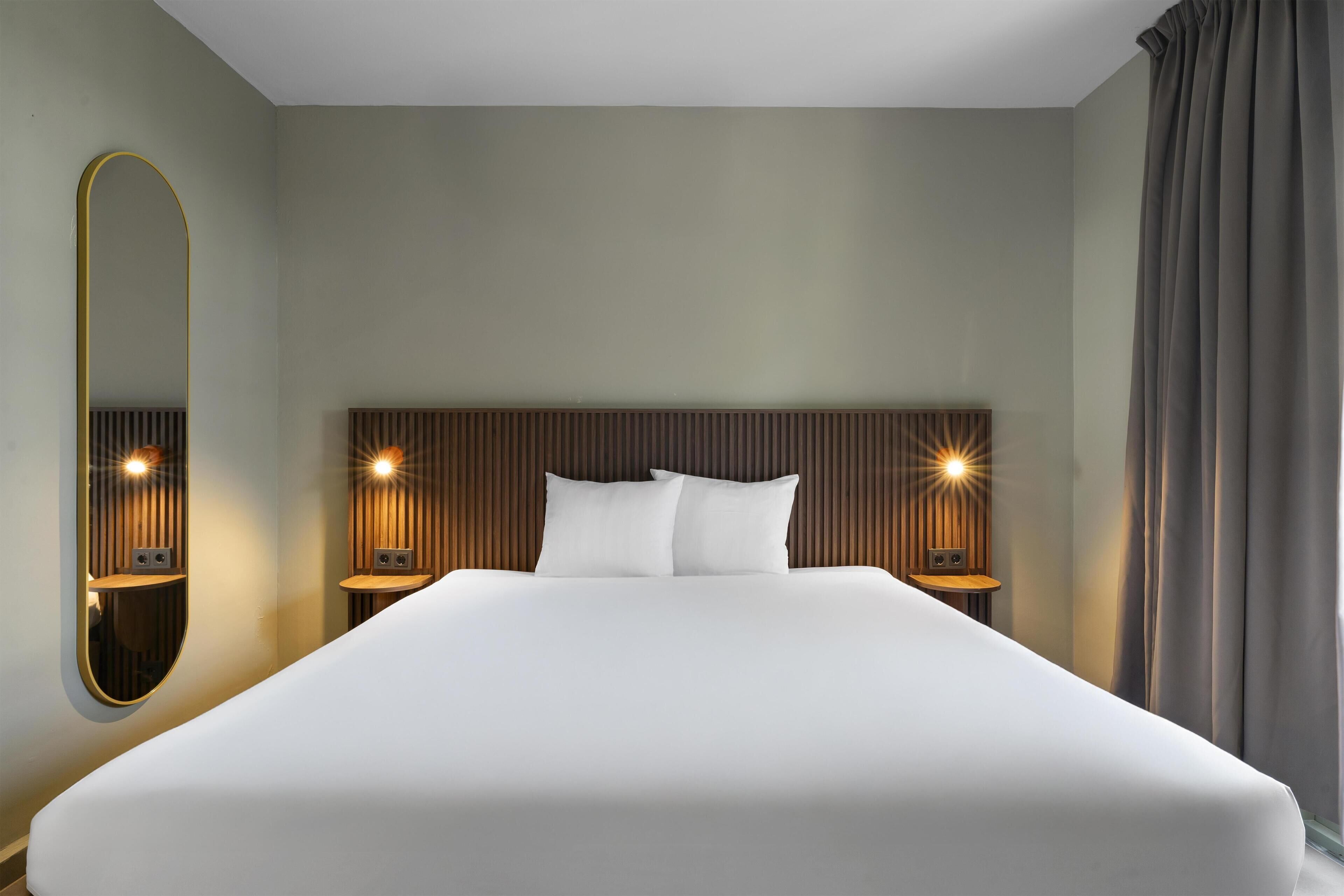 Une chambre d’hôtel avec un grand lit, deux tables de chevet avec des lampes, un miroir et un rideau.