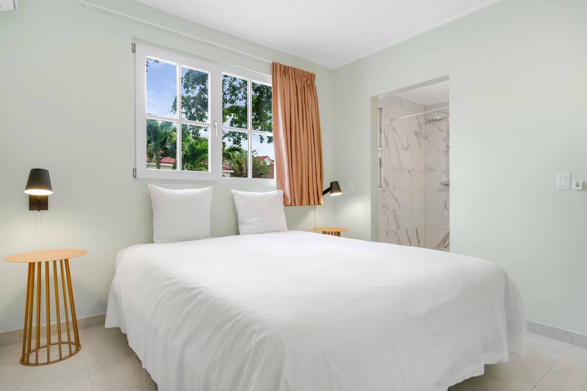 A bedroom with a large bed, a window with curtains, a bedside table, and a lamp.
