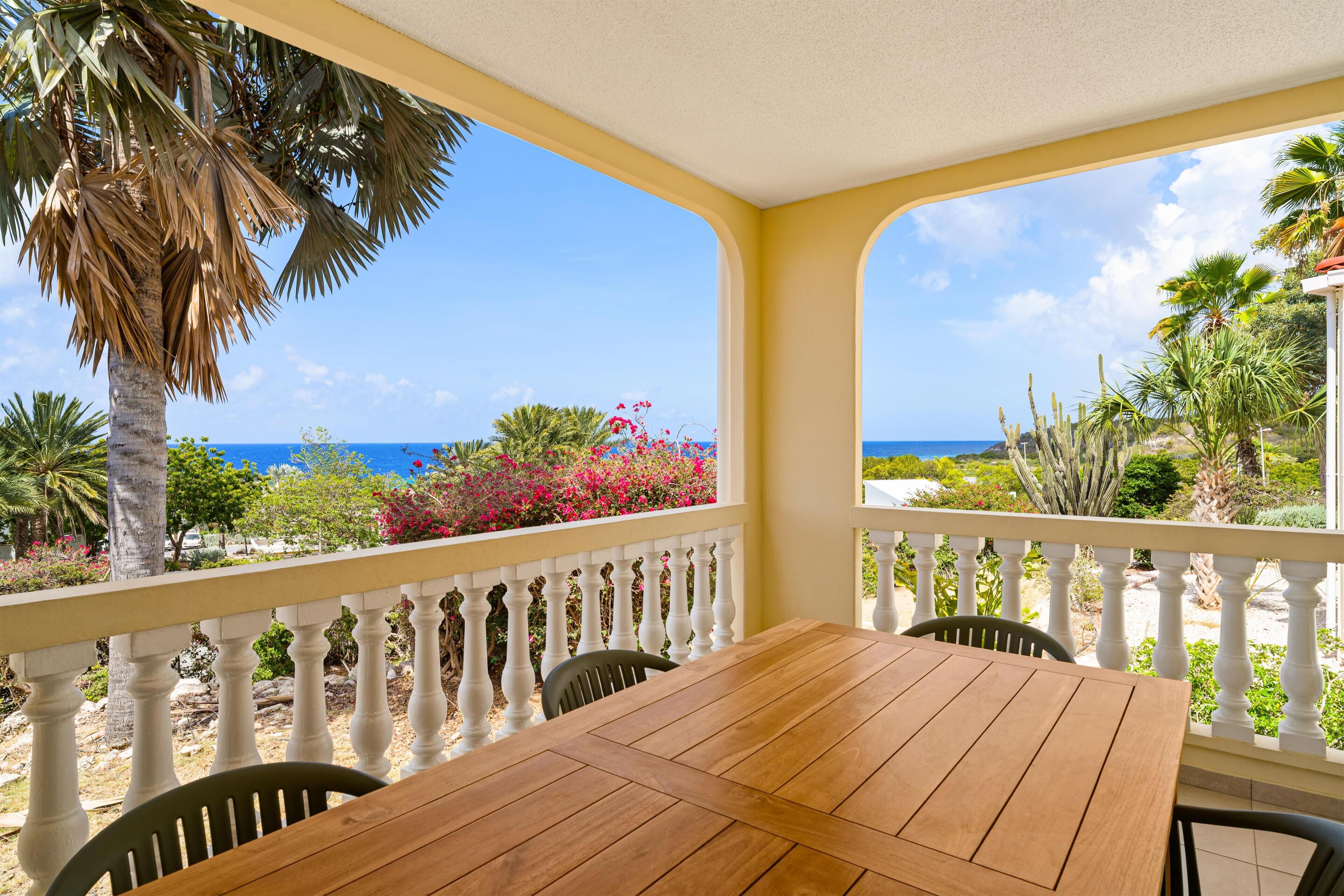 A wooden table and chairs on a balcony with a white balustrade, overlooking a beach with palm trees and cacti.