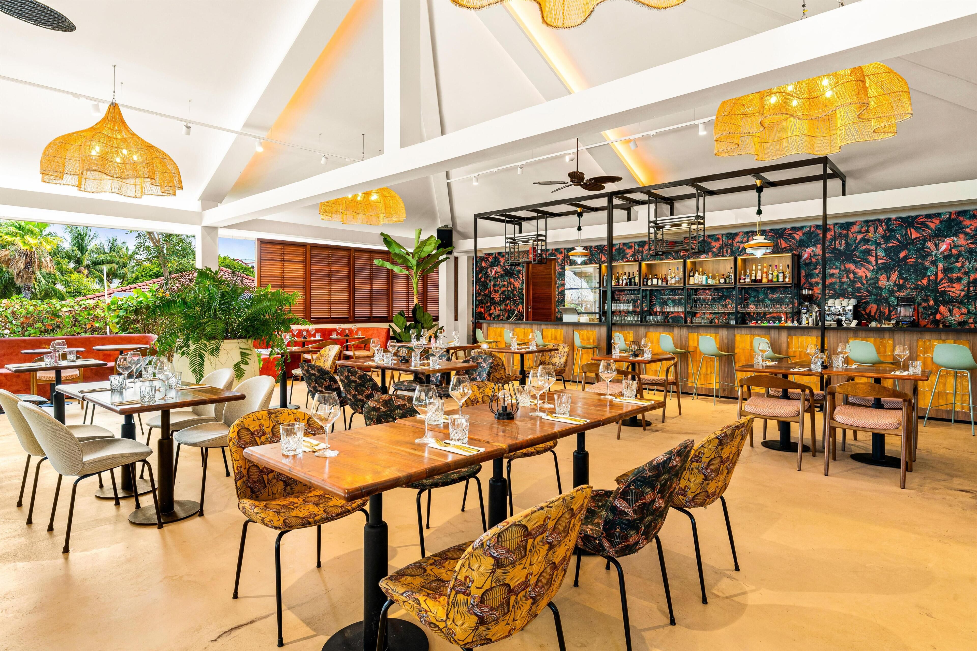 Un restaurante con varias mesas y sillas, una zona de bar con estantes llenos de botellas y vistas a un paisaje tropical.