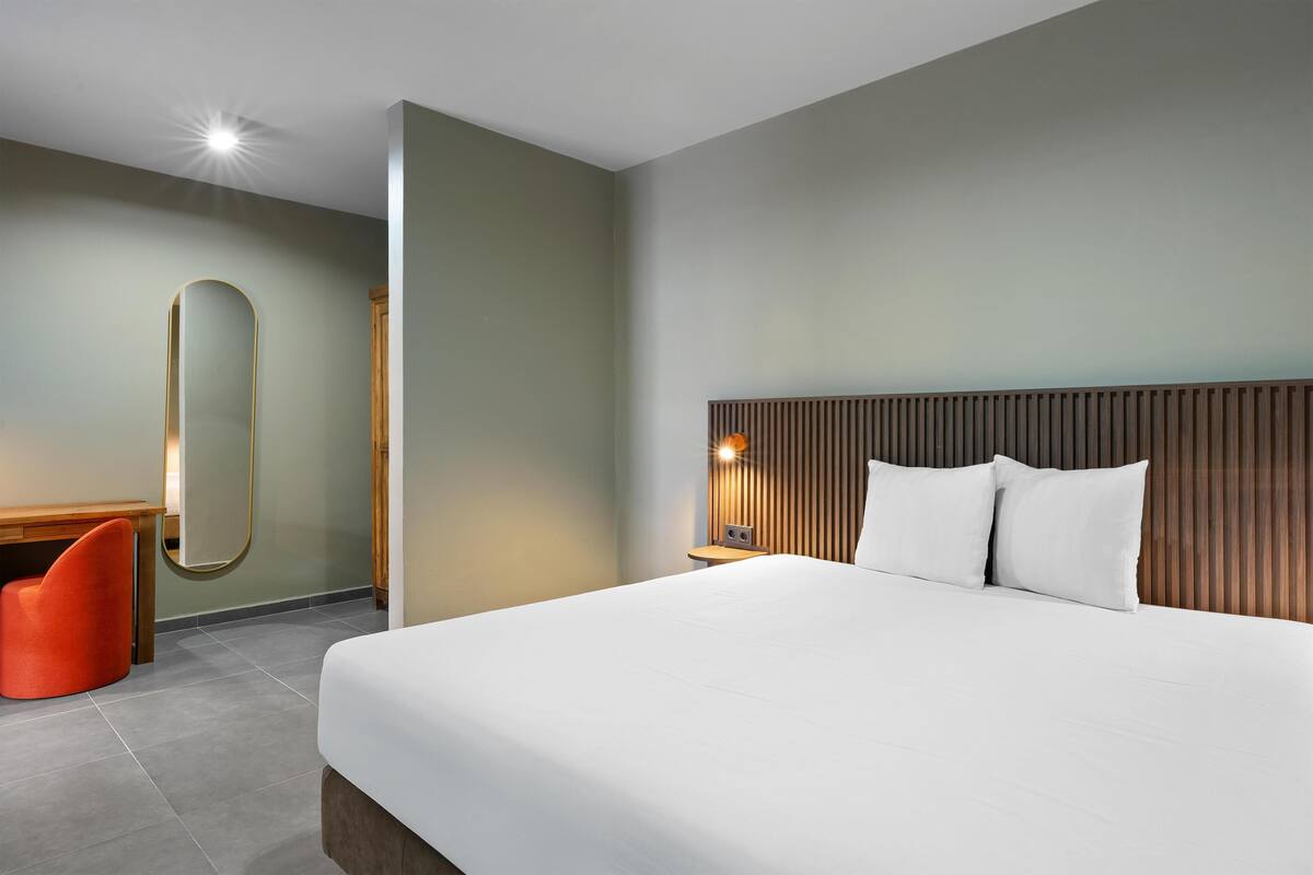 A modern hotel room with a large bed, a wooden headboard, a small desk, and a red chair.
