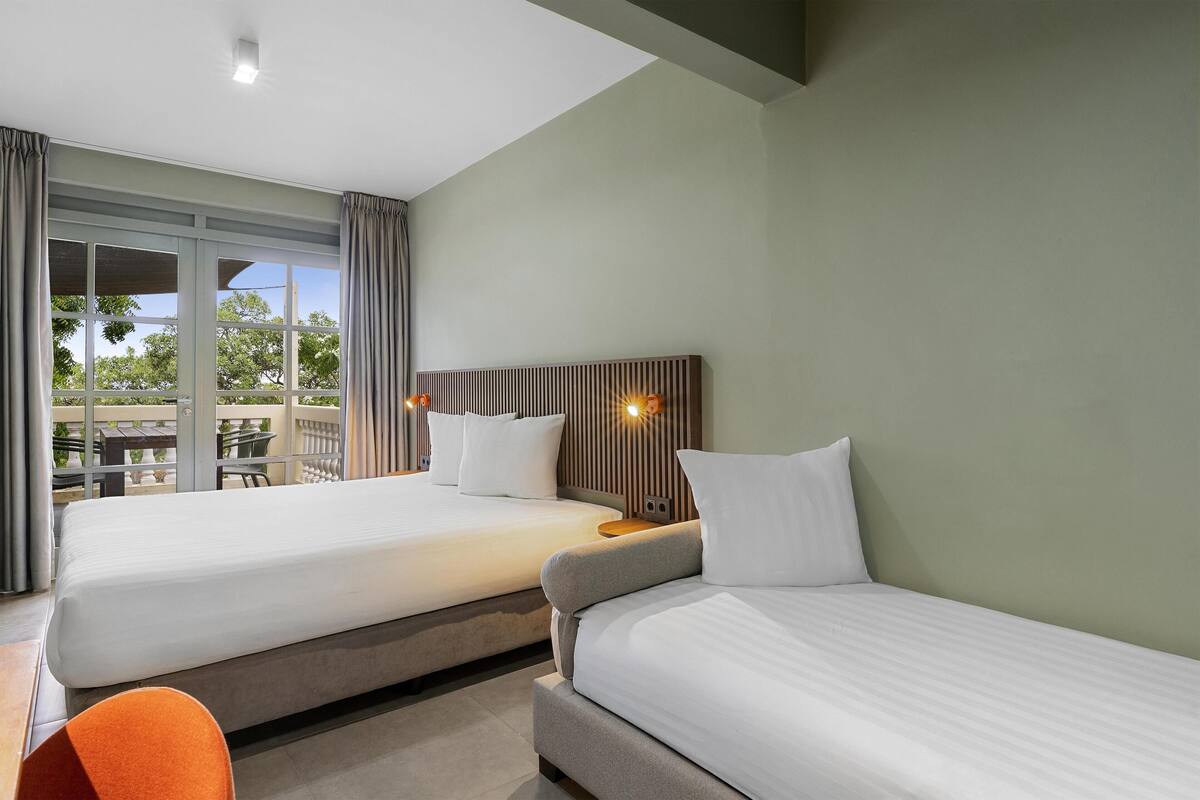 A hotel room with two beds, a large window with curtains, a view of the outdoors, and a small table with a chair.