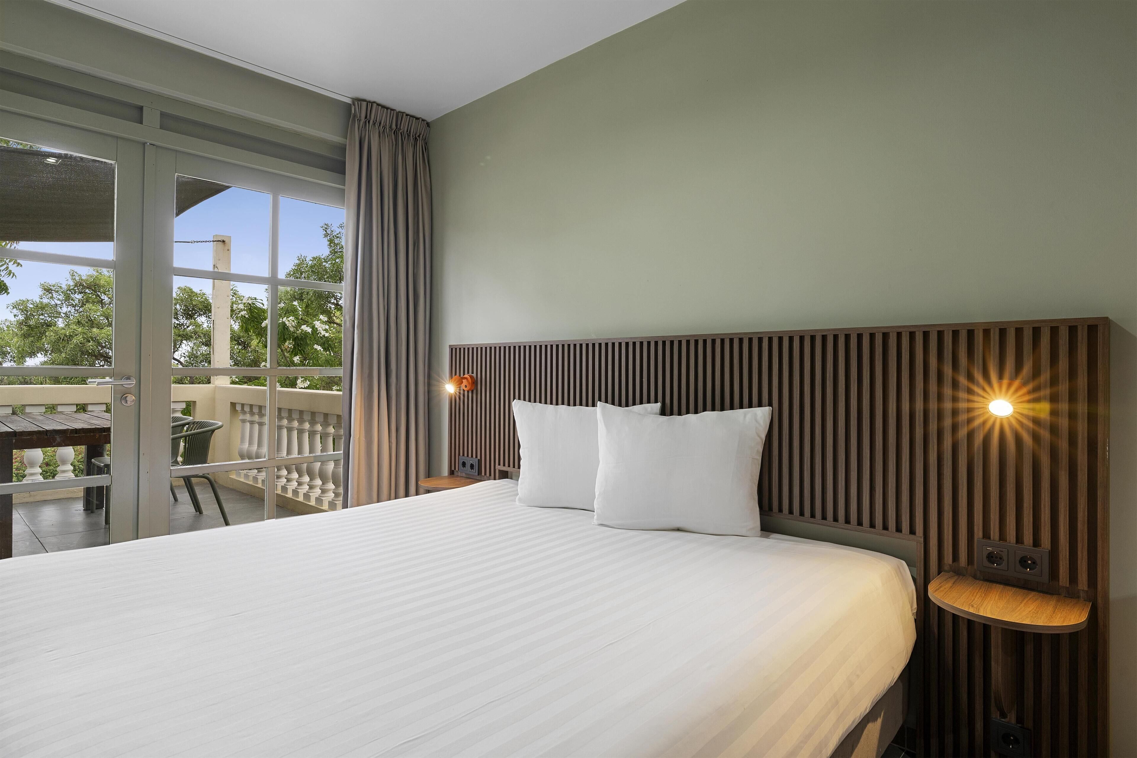 A hotel room with a large bed, a wooden headboard, a small bedside table, and a view of the outdoors.