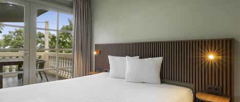 A hotel room with a large bed, a wooden headboard, a small bedside table, and a view of the outdoors.