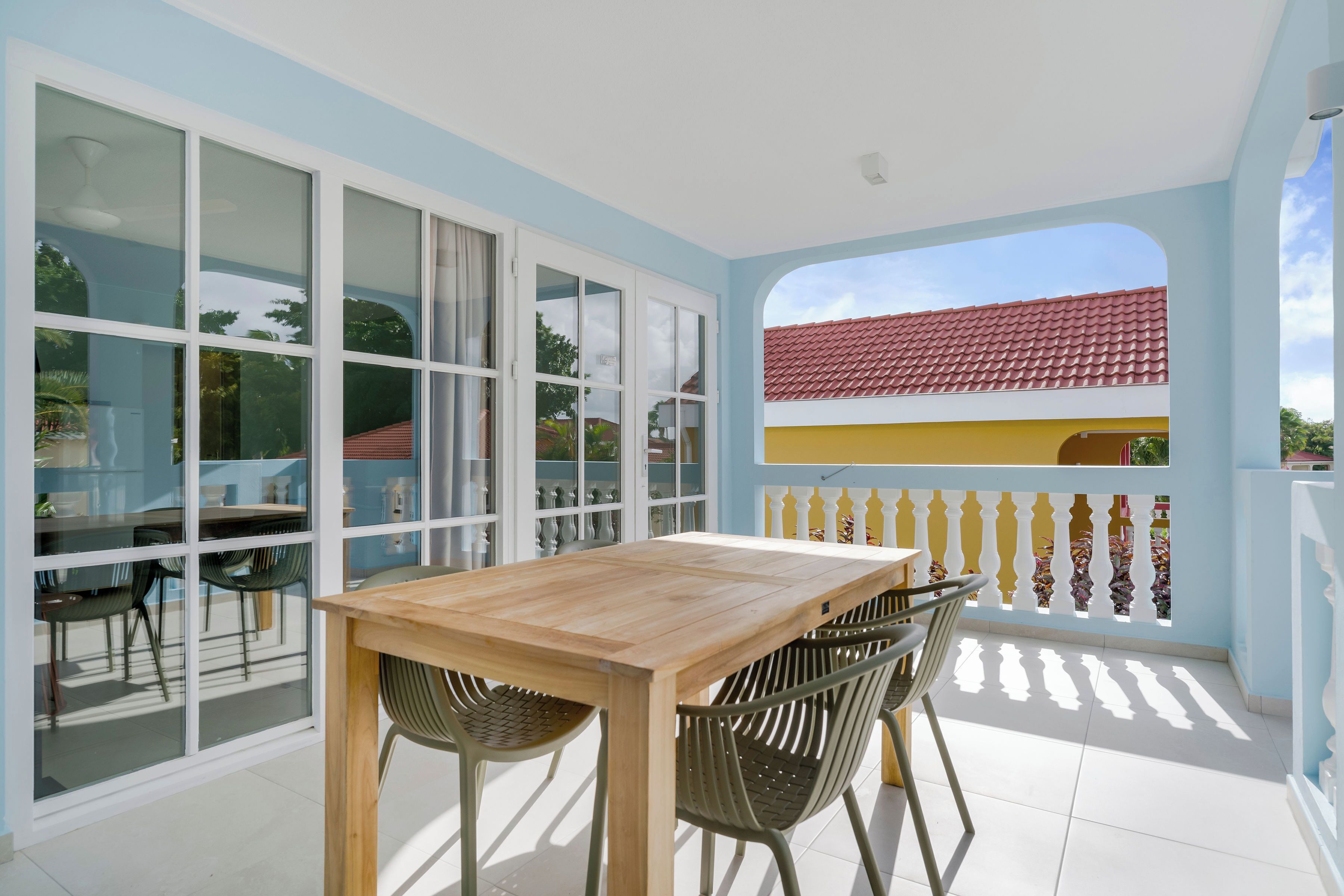 Un balcone con tavolo e sedie in legno, ampie porte in vetro e vista su un tetto con tegole rosse.