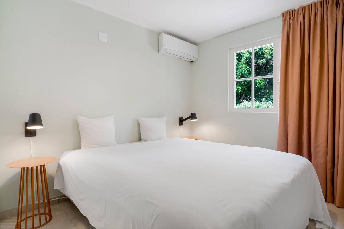 A hotel room with a large bed, two bedside tables, a wall-mounted air conditioner, a window with curtains, and a small round table.