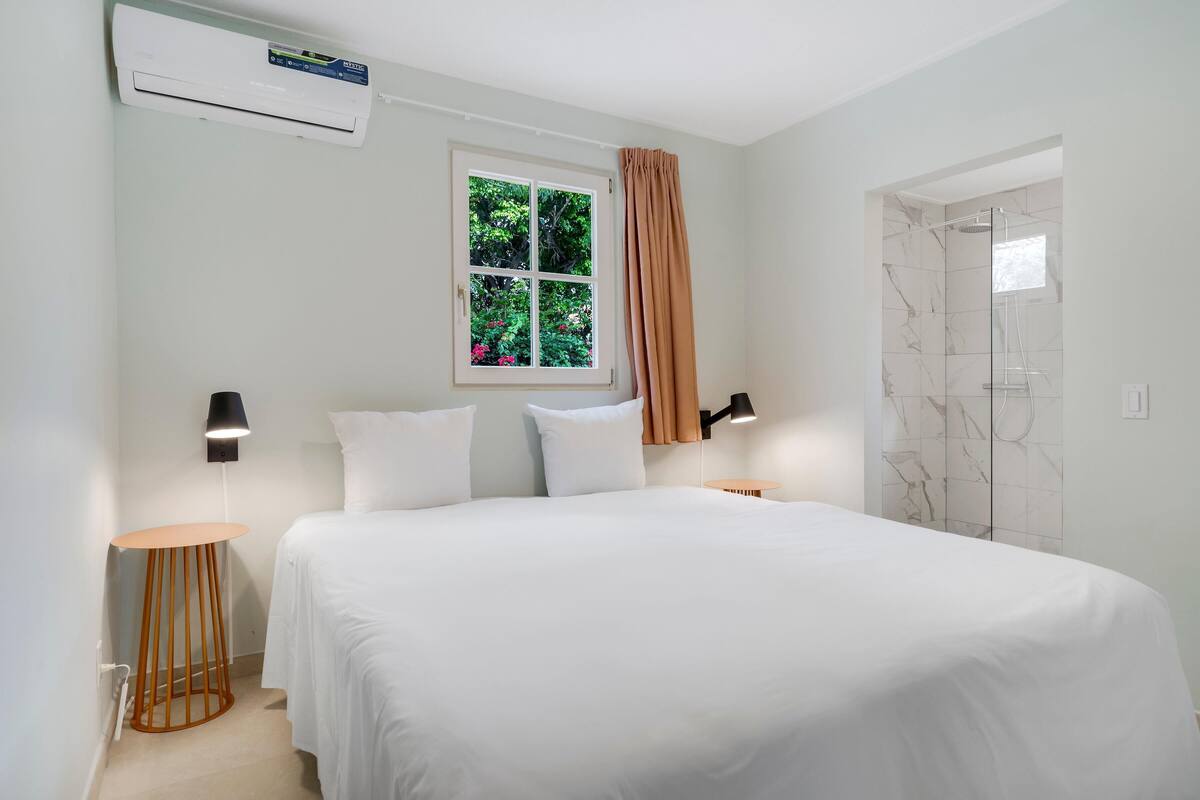 A bedroom with a bed, a window with curtains, a standing lamp, a small round table, and a shower area with a glass door.