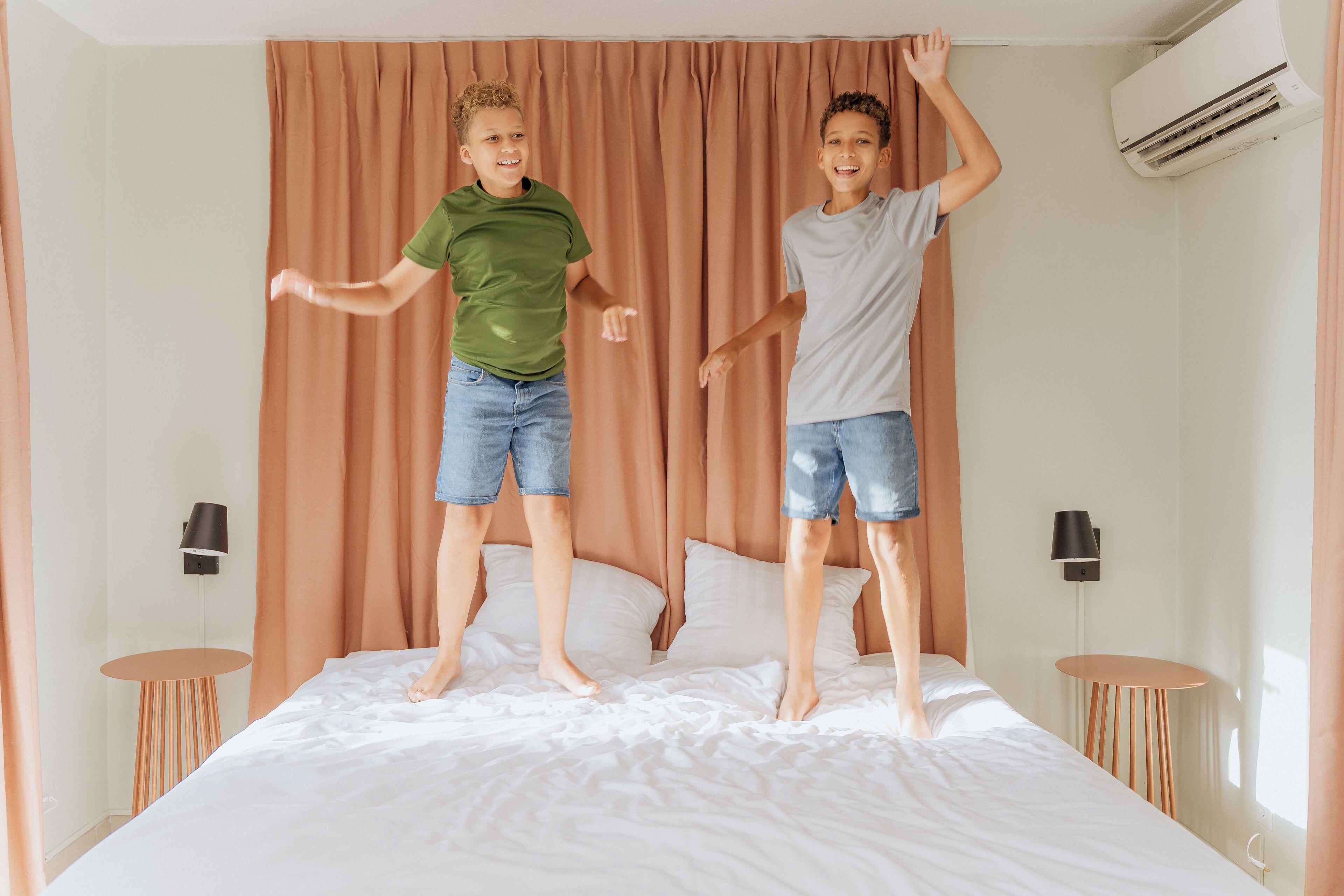 Dos niños saltando sobre una cama en una habitación con cortina, lámpara y aire acondicionado.