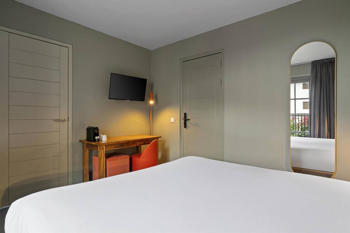 A hotel room with a bed, a desk with a chair, a television mounted on the wall, and a door with a window view.