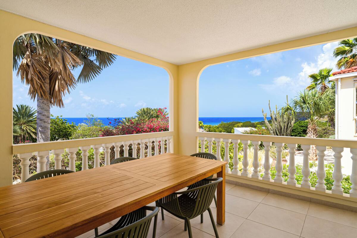A wooden table with chairs on a balcony with white balustrades, overlooking a tropical landscape with palm trees and a view of the ocean.