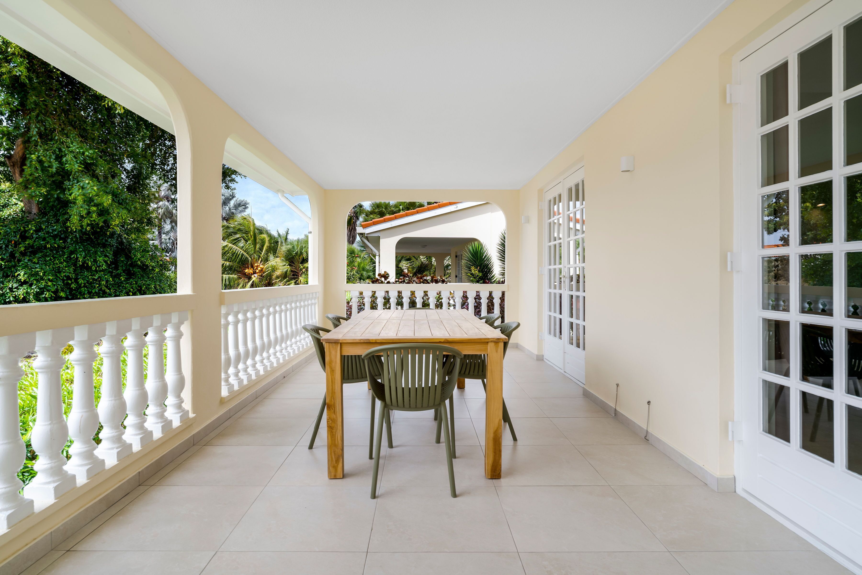 A covered outdoor dining area with a wooden table and chairs, surrounded by white railings and large windows.