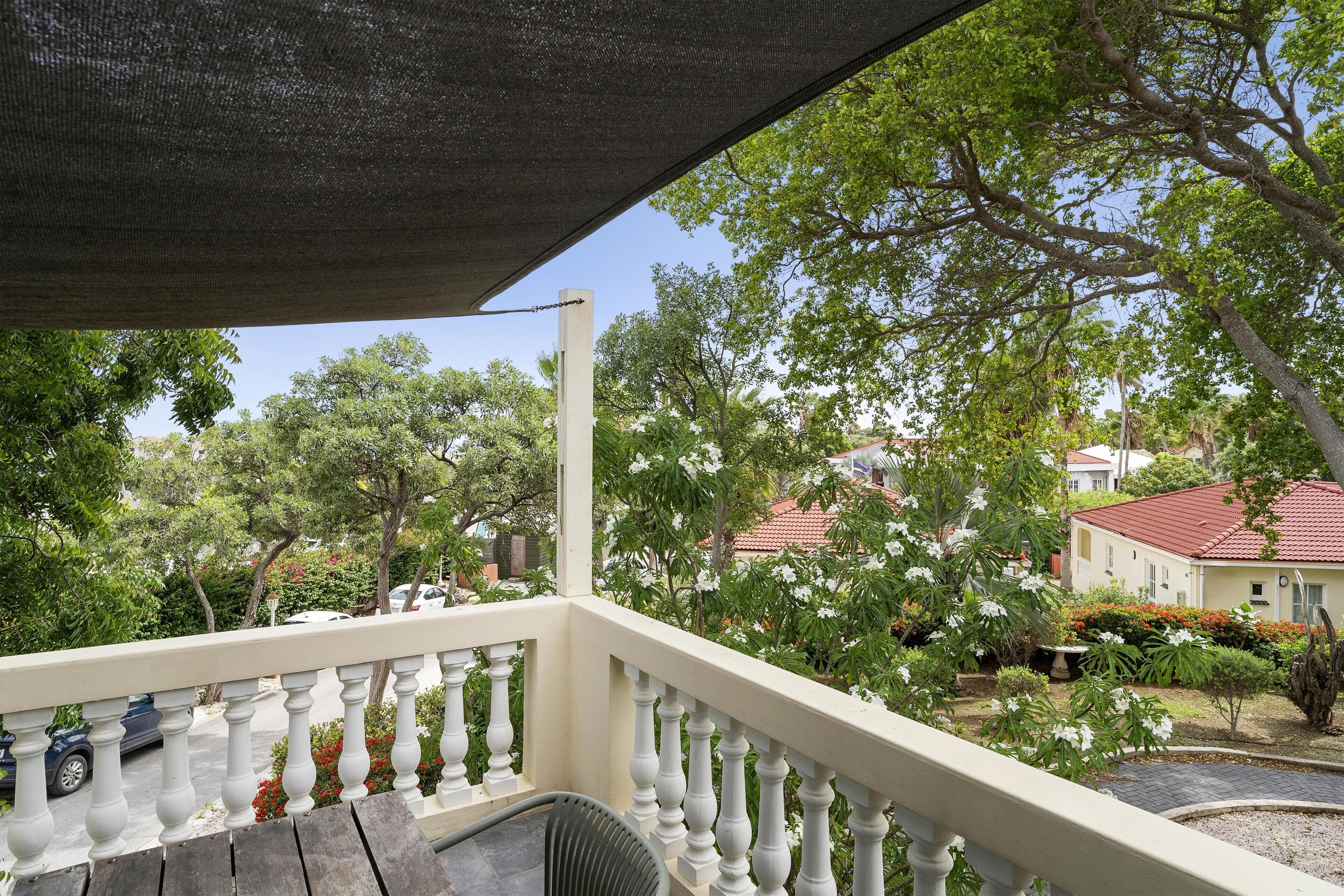 A balcony with a white balustrade, a covered area with a mesh roof, and a view of a residential area with trees and houses.