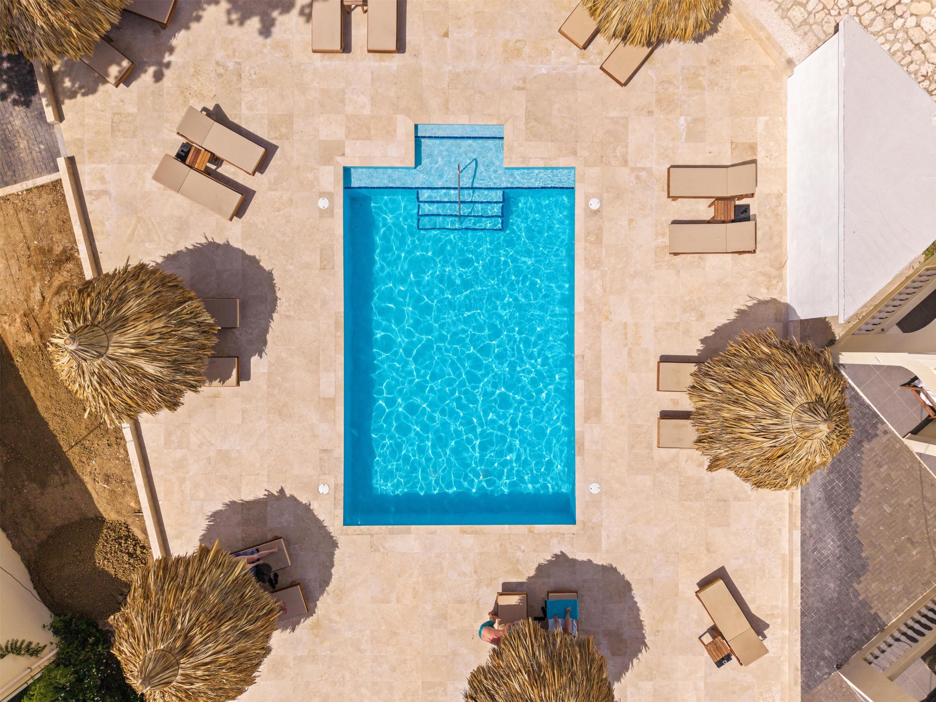 An aerial view of a swimming pool surrounded by lounge chairs and umbrellas, with a stone floor and a clear blue pool.