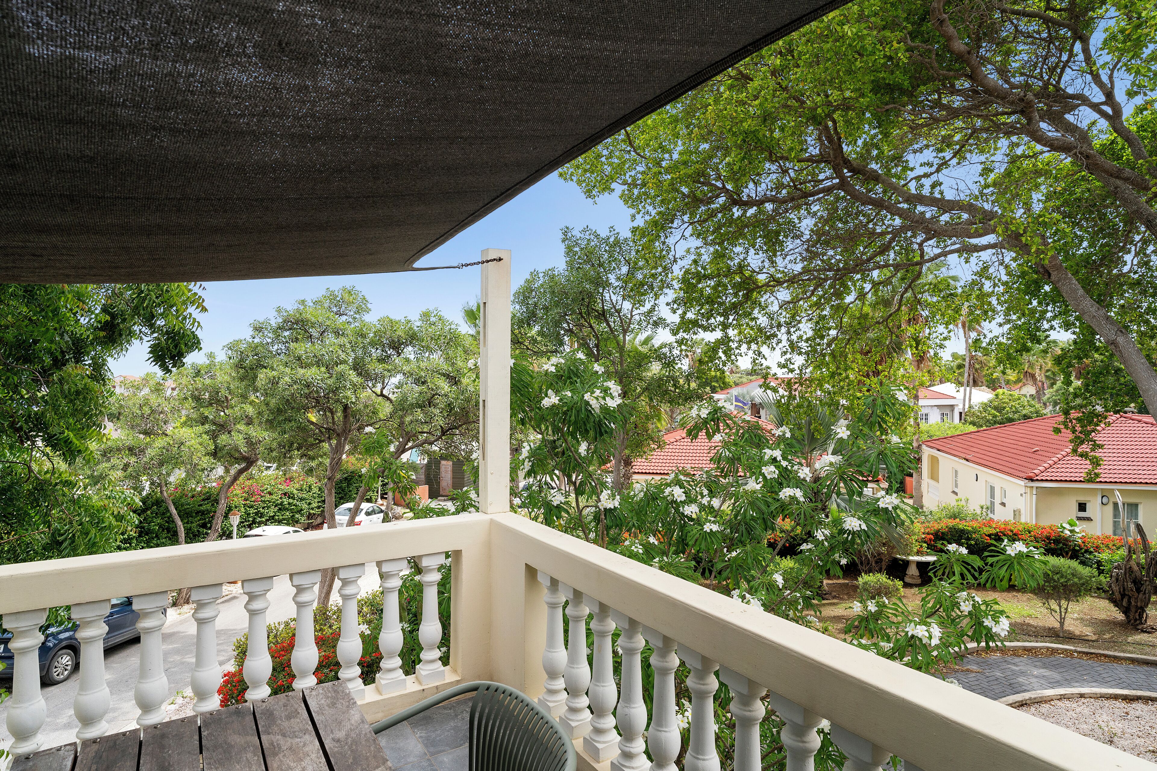 Een balkon met een witte balustrade, met uitzicht op een tuin vol planten en een gebouw met een rood dak op de achtergrond.