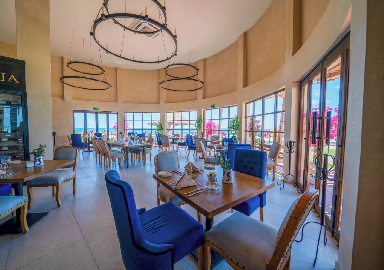 A spacious dining area with multiple tables and chairs, large windows offering views of the outdoors, and a modern chandelier.