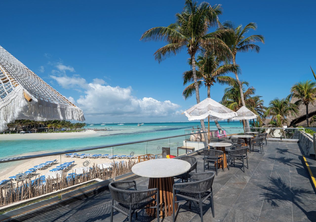 A beachside terrace with tables and chairs, a pool, and palm trees.