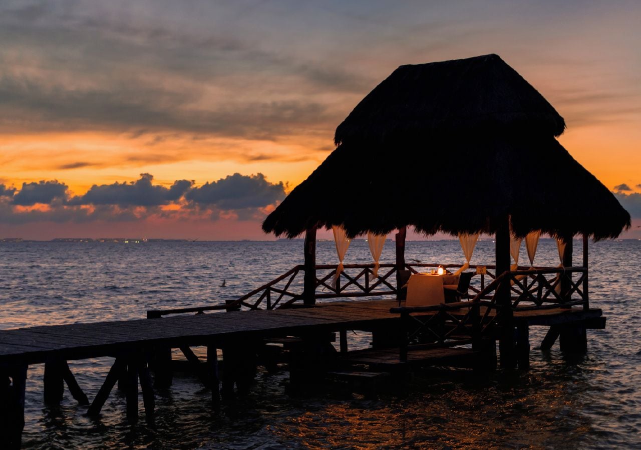 Un muelle de madera que conduce a una construcción con techo de paja y una mesa iluminada con velas, con vista a un cuerpo de agua al atardecer.