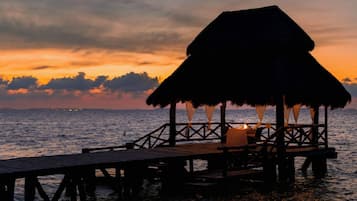 Un muelle de madera que conduce a una construcción con techo de paja y una mesa iluminada con velas, con vista a un cuerpo de agua al atardecer.