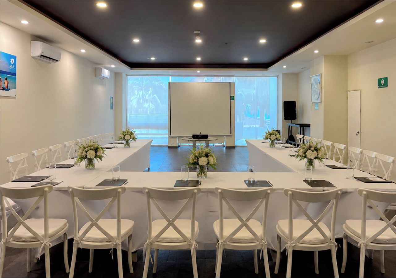 Uma sala de conferências com uma mesa comprida, cadeiras brancas e uma tela de projetor.
