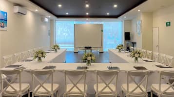 Uma sala de conferências com uma mesa comprida, cadeiras brancas e uma tela de projetor.