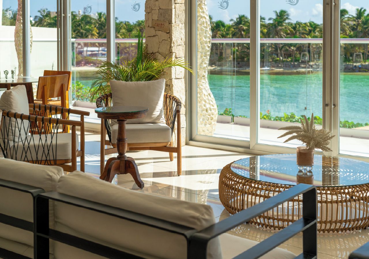 A sunlit lounge area with a view of a turquoise sea, featuring wicker furniture and a glass table.