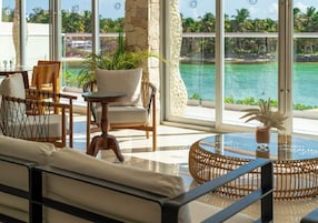 Un área de estar soleada con vista a un mar turquesa, que incluye muebles de mimbre y una mesa de vidrio.