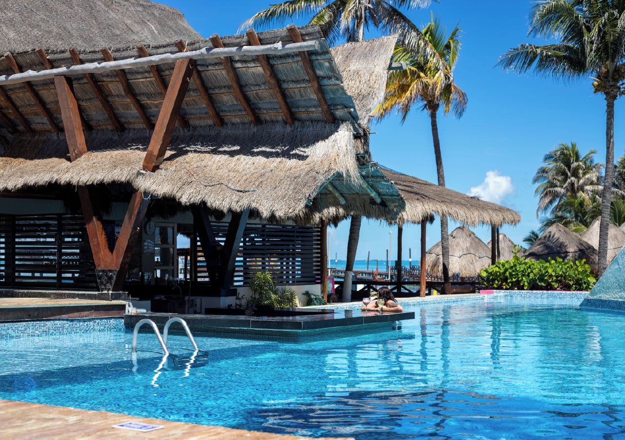 A swimming pool with a thatched-roof structure and palm trees in the background.