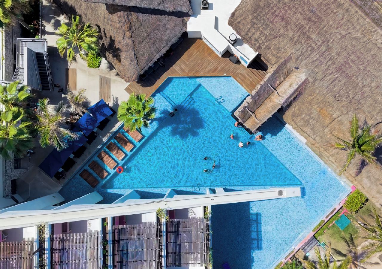 An aerial view of a hotel with a large swimming pool, multiple lounge areas, and palm trees.