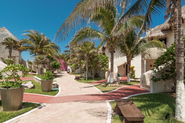A resort courtyard with palm trees, a paved walkway, and thatched-roof buildings. - Mia Reef Isla Mujeres - All Inclusive (Isla Mujeres)