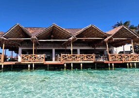Bungalows sobre el agua con techos de paja y barandillas de madera, unidos por una pasarela de madera sobre aguas turquesas cristalinas.