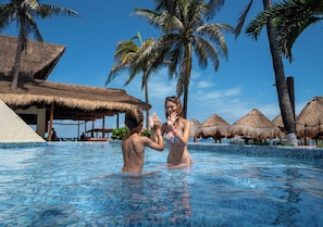 A swimming pool with palm trees and thatched-roof structures in the background. - Mia Reef Isla Mujeres - All Inclusive (Isla Mujeres)