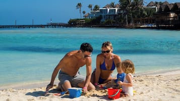 Escena de playa con mar azul turquesa, un muelle al fondo y un resort con bungalows sobre el agua.