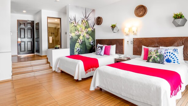 A hotel room with two beds, wooden flooring, a large painting, a wooden headboard, wall-mounted lights, and a potted plant. - Mia Reef Isla Mujeres - All Inclusive (Isla Mujeres)