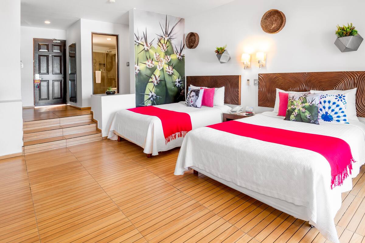 Ein Hotelzimmer mit zwei Betten, Holzboden, einem großen Gemälde, einem hölzernen Kopfteil, Wandleuchten und einem Blumentopf.