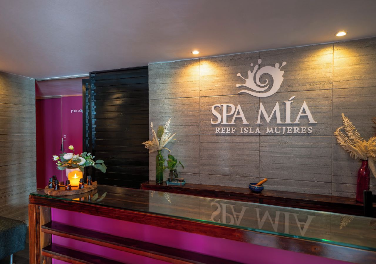 A reception area with a wooden reception desk, a wall-mounted sign reading 'SPA MIÁ REEF ISLA MUJERES', and a small plant on the desk.