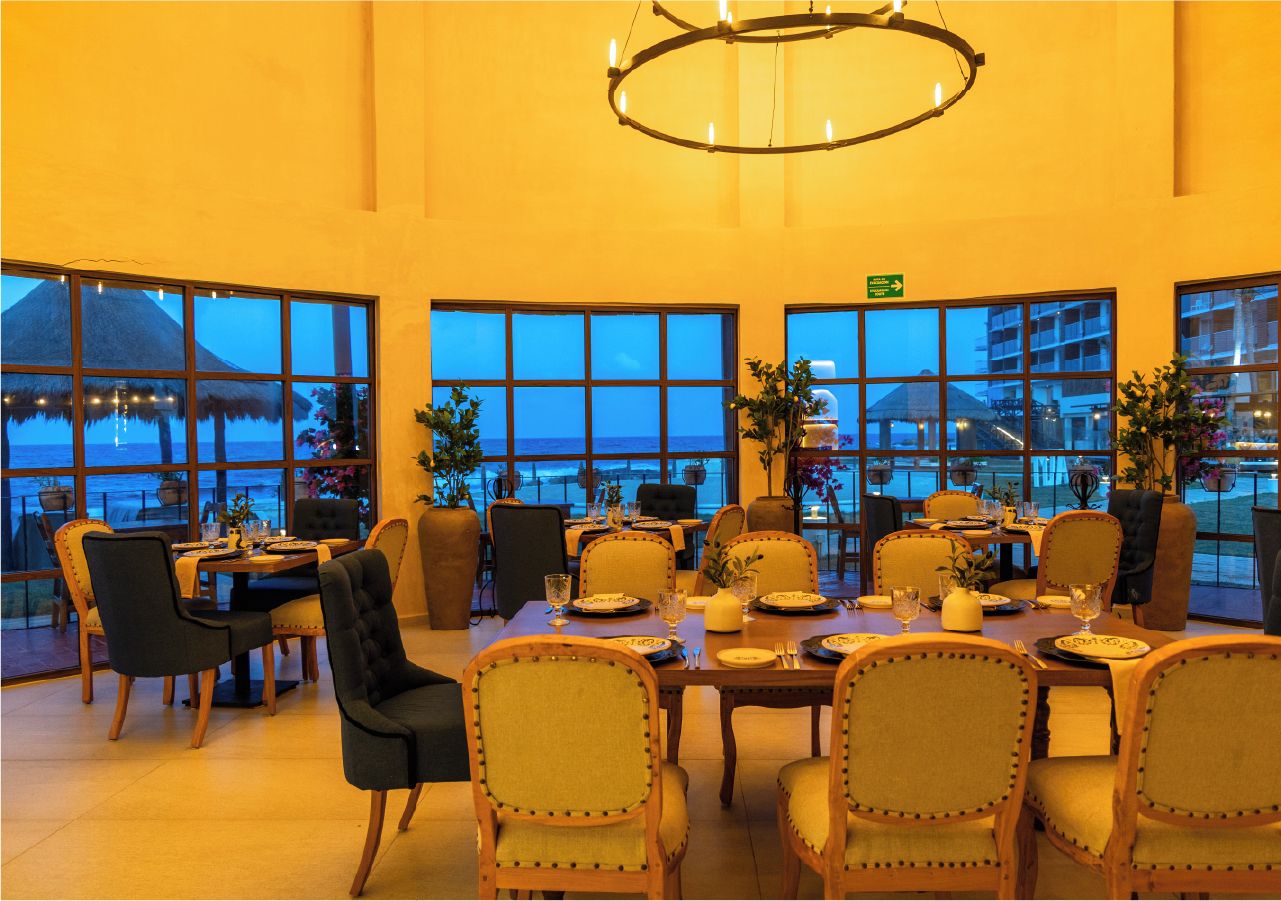 A dining area with tables set for guests, large windows offering a view of the sea, and a chandelier hanging from the ceiling.