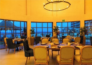 A dining area with tables set for guests, large windows offering a view of the sea, and a chandelier hanging from the ceiling. - Mia Reef Isla Mujeres - All Inclusive (Isla Mujeres)