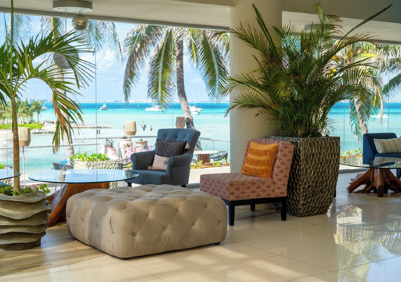 Un área de descanso moderna con vista al océano, que incluye un otoman capitonado, un sillón azul y otro sillón estampado.