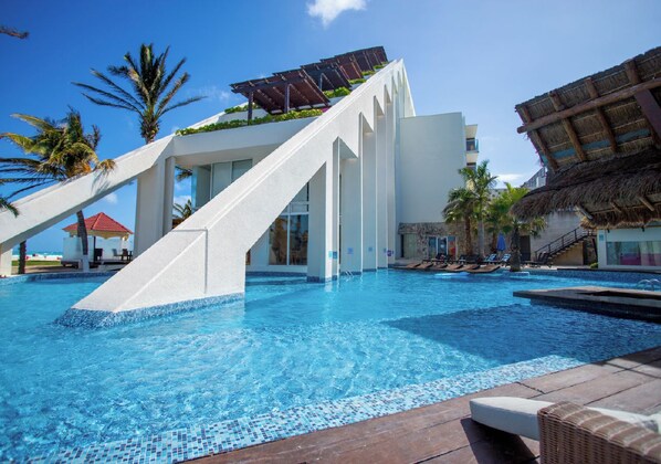 A swimming pool with a white structure, palm trees, and a thatched roof. - Mia Reef Isla Mujeres - All Inclusive (Isla Mujeres)