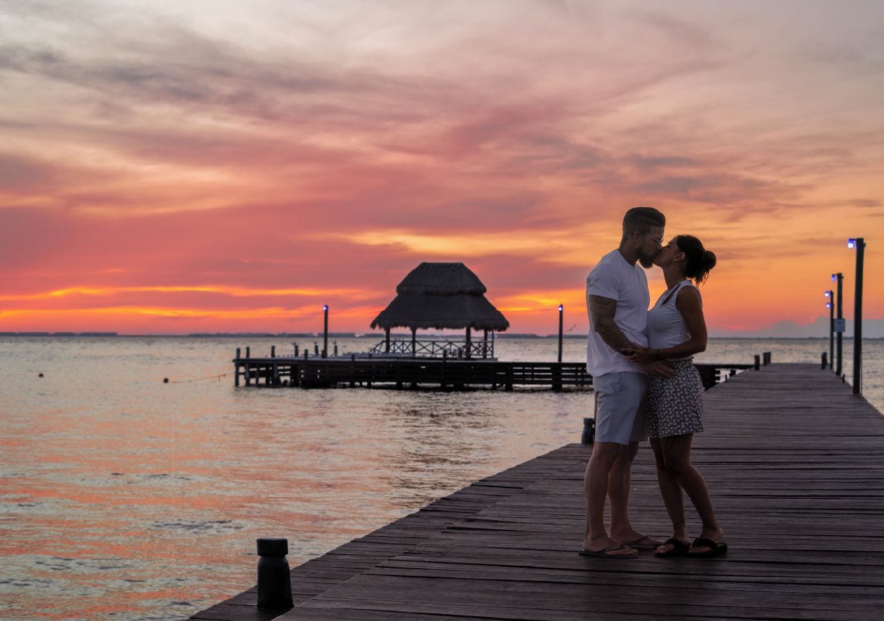 A couple standing on a wooden pier overlooking a body of water with a thatched-roof structure in the distance.