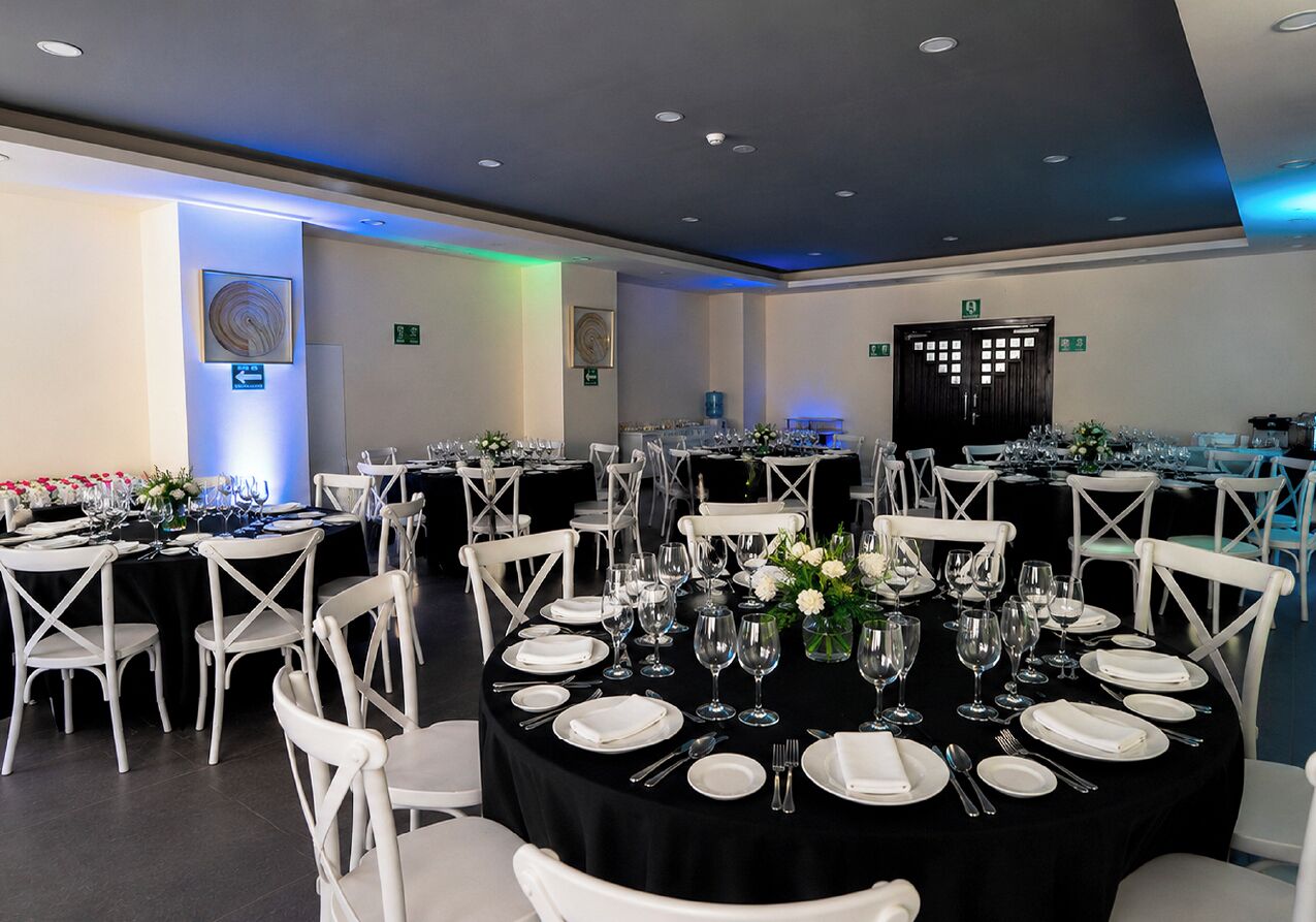 Un salón de banquetes con mesas redondas dispuestas para cenar, sillas blancas y mantel negro.