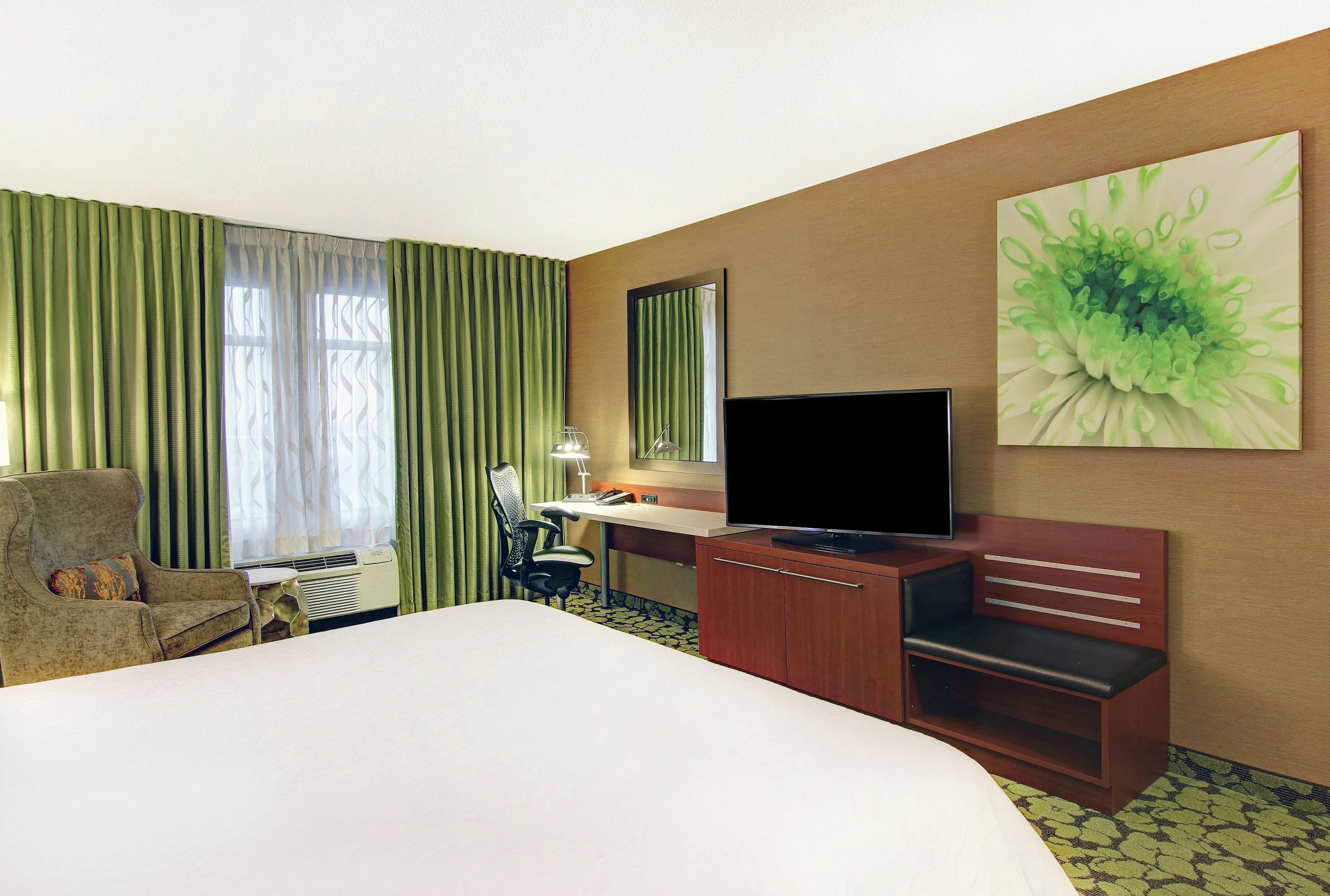 A hotel room with a bed, a desk with a computer, a television, a chair, and a large window with curtains.
