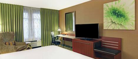 A hotel room with a bed, a desk with a computer, a television, a chair, and a large window with curtains.