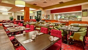 A hotel dining area with multiple tables and chairs, a kitchen in the background, and a red and green color scheme.