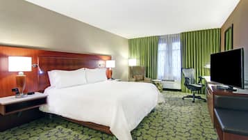 A hotel room with a large bed, two bedside tables with lamps, a desk with a flat-screen TV, a chair, and a large window with green curtains.