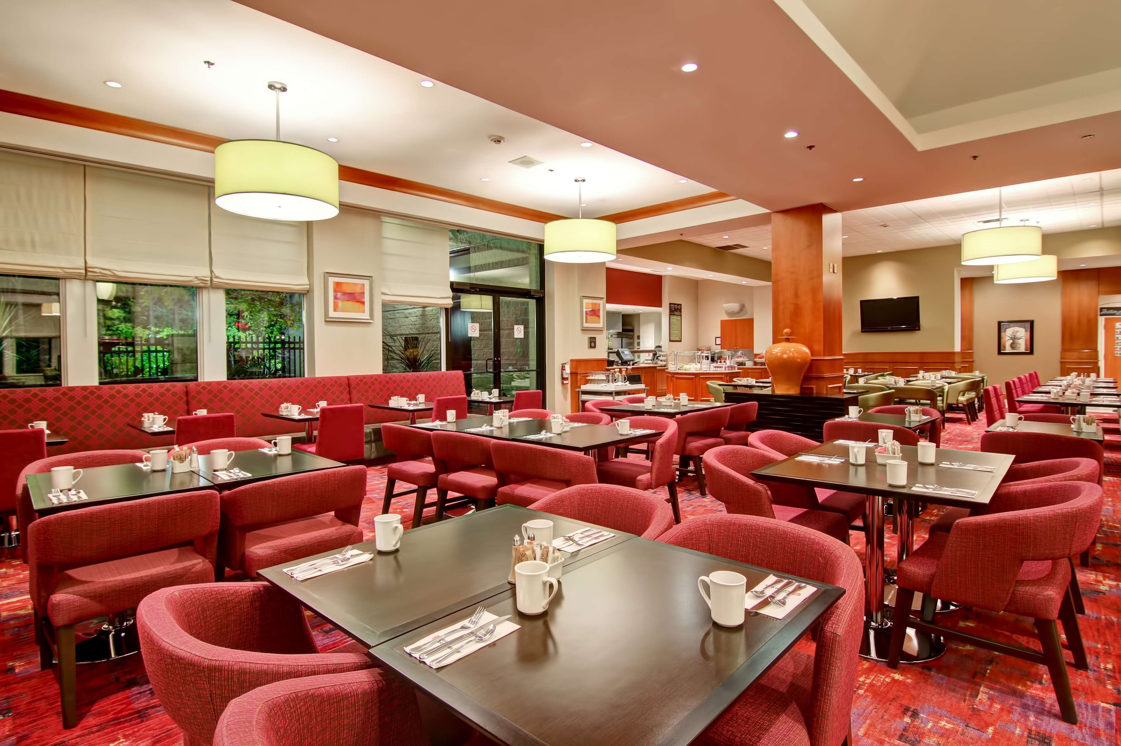 A hotel dining area with red upholstered booths and tables, large windows, and a kitchen in the background.