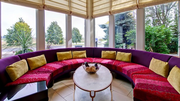 A curved purple sofa with gold cushions, a round table with a basket, and large windows with a view of greenery. - Hilton Garden Inn Toronto/Markham (Markham)