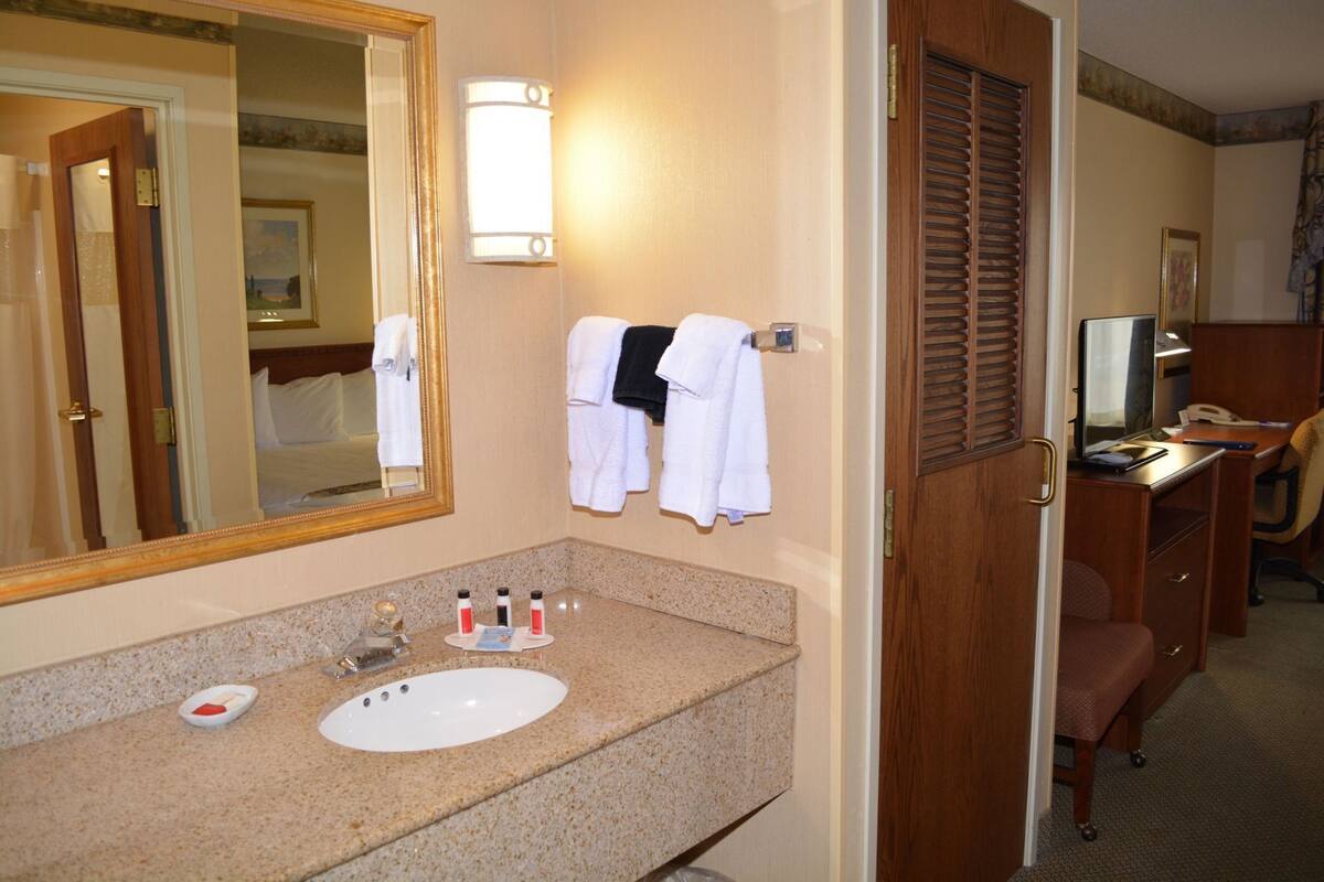 Un baño de hotel con lavamanos, espejo, toallero y un escritorio con una silla.